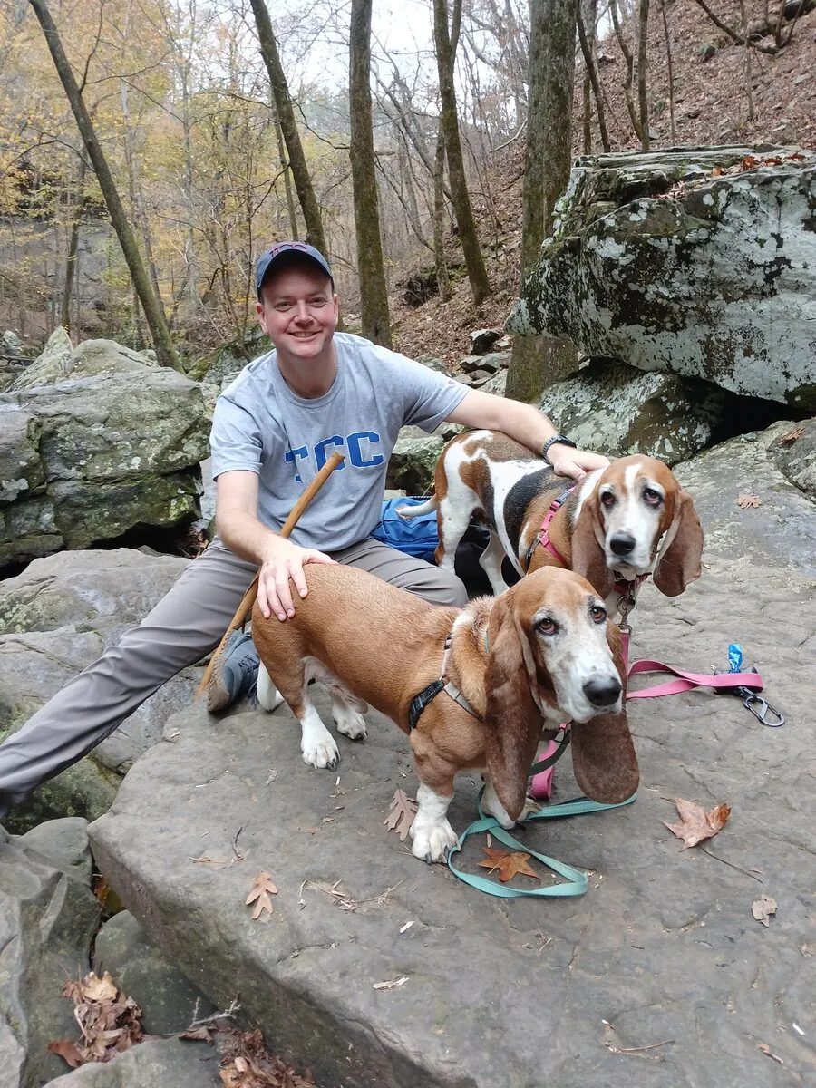 Jared in a TCC shirt crouched on a trail boulder with his two basset hounds